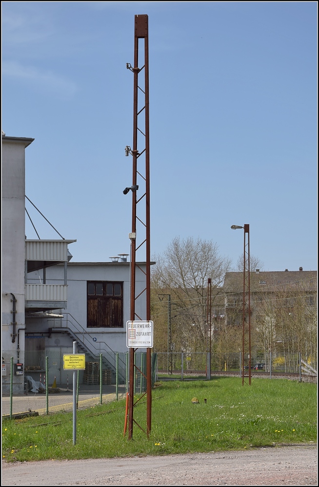 Anschlussverwendung der Oberleitungsmasten der badischen Staatsbahn in Lörrach-Brombach an einem ehemaligen Werksgleis. Kamerainstallation und Lampenmast. April 2013

Die ersten ernstzunehmenden Elektrifierzierungen mit Einphasenwechselstrom wurden anfangs mit 15 kV und 15 Hz durchgeführt. Durch einen Beschluss der badischen, bayrischen und hessisch-preußischen Staatsbahnen im Jahr 1912 wurden 16 2/3 Hz festgelegt. Die BLS schloss sich dem an und stellte ihre bereits elektrifizierte Strecke gleich darauf um, die RhB verwendete ebenso 16 2/3 aber eine reduzierte Spannung von 11 kV, die SBB hingegen begann erst ab 1919 mit Elektrifizierungen. In Deutschland waren Wiesen- und Wehratalbahn das größte elektrifizierte Netz ab 1913, immerhin bis 1936 mit 15 Hz. Der 1. Weltkrieg bremste erst einmal weitere Elektrifizierungen bis 1922 aus. Somit dürften diese Masten 105 Jahre alt sein. Kurz vor Zell im Wiesental sind sogar noch einige dieser Masten in Betrieb. Im Bild der vierte Mast ist aus der Reichsbahnzeit.