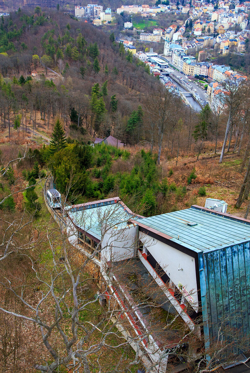 Blick auf die Bergstation der Standseilbahn Diana (tschechisch Lanová dráha Diana) und im Tal aus Karlsbad (Karlovy Vary), vom 40 m hohen Diana-Aussichtsturm auf der Freundschaftshöhe 556 m ü.M. am 18.04.2023. Der Wagen „DIANA 1“ der Standseilbahn fährt gerade wieder hinab.

Die Stadtseilbahnen (Diana sowie Imperial) stellen in Karlsbad schon seit Anfang des vergangenen Jahrhunderts ein traditionelles und beliebtes Verkehrsmittel dar. Sie gehören inzwischen zum Stadtbild und sind ein beliebtes Touristenziel.

Diana-Standseilbahn:
Die Standseilbahn Diana (tschechisch Lanová dráha Diana) wurde im August 1912 eröffnet und beginnt an der Station Alte Wiese (Stará louka) auf einer Höhe von 389 m und führt auf die Freundschaftshöhe 556 m über NN, wo sich der Diana-Aussichtsturm befindet. Die Talstation befindet sich gleich neben dem Grandhotel Pupp in der Gasse Mariánská. Den Höhenunterschied von 167 m überwindet sie auf einer Strecke von 437 m. Im Bereich der abtsche Ausweiche liegt auch die Mittelstation Jelení skok (Hirschsprung) in 473 m Höhe. Die maximale Steigung beträgt 432,3 ‰, durchschnittlich liegt diese bei 398,1 ‰. Die Bahn wurde insgesamt dreimal renoviert, zwischen 1963 und 1965, 1972 und noch einmal zwischen 1984 und 1988. Im Gegensatz zur Bahn zum Personennahverkehrs der Stadt. Auch sie wird im 15-Minuten-Takt befahren und benötigt 4 Minuten für eine Fahrt bei einer Geschwindigkeit von 2,04 m/s. Die Gleise sind vom Typ A, die Spurweite beträgt 1.000 mm. Die leer 7.900 kg wiegenden Wagen sind für 49 + 1 Fahrer ausgelegt. Das Seil hat einen Durchmesser von 28 mm. Die Antriebsleistung beträgt 51 kW, die Antriebsregulation erfolgt mit Hilfe von Thyristoren, 

Die Seilbahn ist ein Projekt des Ing. H. H. Peter aus Zürich. Der Bau der Seilbahn wurde von der Wiener Baufirma Leo Arnoldi mit Hilfe von lokalen Handwerkern und Firmen durchgeführt. Die erste Generalüberholung der Seilbahn geschah in den Jahren 1963-1965, als die Seilbahnen neue Metallwagons aus der Produktion des Tatra Smíchov Wagon bekamen. Die Rekonstruktion der elektrischen Ausrüstung wurde 1972-73 durchgeführt.Es ist ein traditionelles und sehr beliebtes Verkehrsmittel auf der Freundschaftshöhe in Karlsbad, das hauptsächlich von den Touristen der Stadt genutzt wird. Von der Talstation wird man zum Gipfel gebracht, wo sich der Aussichtsturm Diana und das Wald-Restaurant und Café Diana befindet.

Betreiber der Seilbahnen ist die DPKV - Dopravní podnik Karlovy Vary a. s. (Verkehrsbetrieb Karlsbad AG). Der Verkehrsbetrieb Karlsbad AG ist ein traditioneller Betreiber des öffentlichen Verkehrs in Karlsbad. Er betreibt tagsüber und auch in den Nachtstunden die regelmäßigen Stadtbusverkehrslinien, Sonderverkehrslinien und Vorortverkehrslinien. Ein Besonderheit des Karlsbader ÖÖV ist der Verkehr der Stadtseilbahn vom Theaterplatz (Divadelní náměstí) zum Hotel Imperial und vom Grandhotel Pupp zum Aussichtsturm Diana. Der Verkehrsbetrieb Karlsbad AG bietet der Öffentlichkeit auch weitere, überwiegend mit der Beförderung zusammenhängende Dienstleistungen an. 

DATEN der Seilbahn DIANA:
Höhenunterschied: 167 m
Streckenlänge:  453 m
Spurweite: 1000 mm (Meterspur)
Maximale Steigung:  432,3 ‰
Leergewicht eines Wagens: 7.900 kg
Kapazität eines Wagens: 49 + 1 Fahrer
Seildurchmesser: 28 mm
Antriebsleistung: 51 kW
Höchstgeschwindigkeit: 7,3 km/h
Taktung: alle 15 Minuten
Fahrtdauer: ca. 3 Minuten
Preis (z.Z.) für Hin- und Rückfahrt: 150 CZK