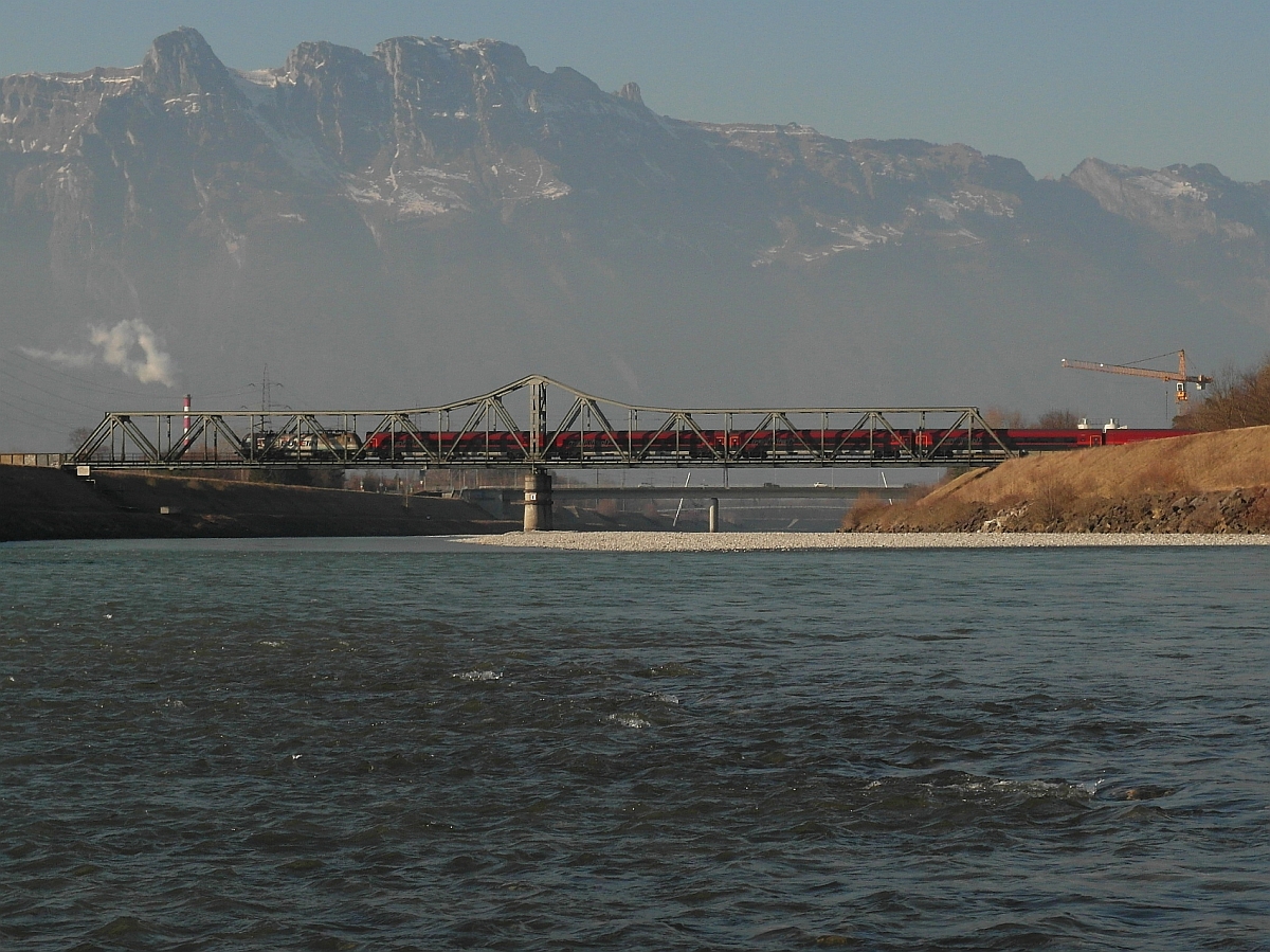 Grenzbild - Schweizer Berge im Hintergrund, schweizer Ufer links, lichtensteiner Ufer rechts und auf der Brücke die österreichische 1116 222 'RED BULLETiN', die den RJ 169, Zürich-Wien, über die Grenze schiebt. Fotostandpunkt: Kiesbank im Rhein bei Buchs am 07.03.2014.