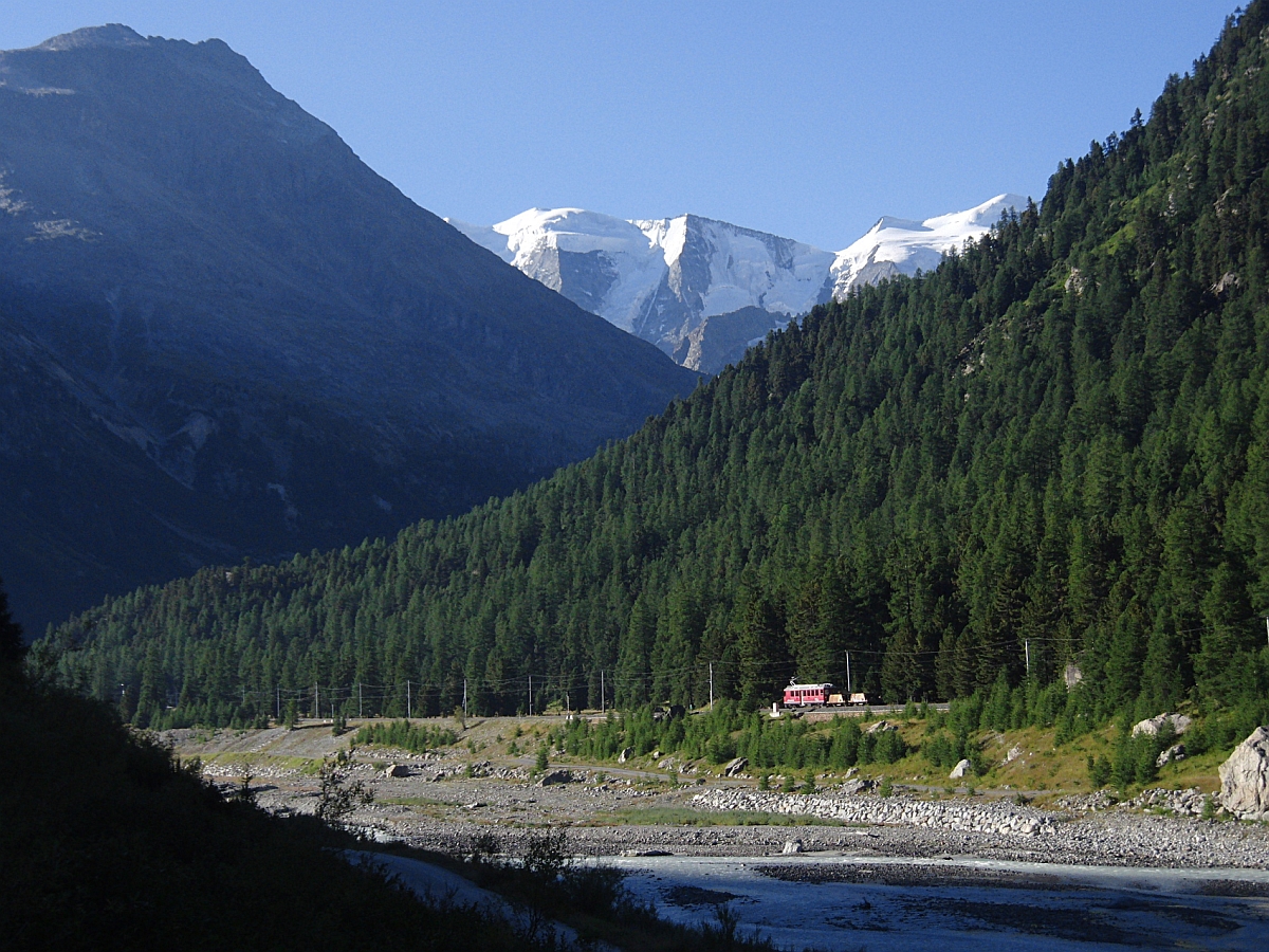 Mit Blick auf die schneebedeckten Berge der Bernina-Gruppe schiebt am 20.08.2012 ein Triebwagen zwei Schotterwagen zwischen Morteratsch und Surovas talwärts Richtung Pontresina.