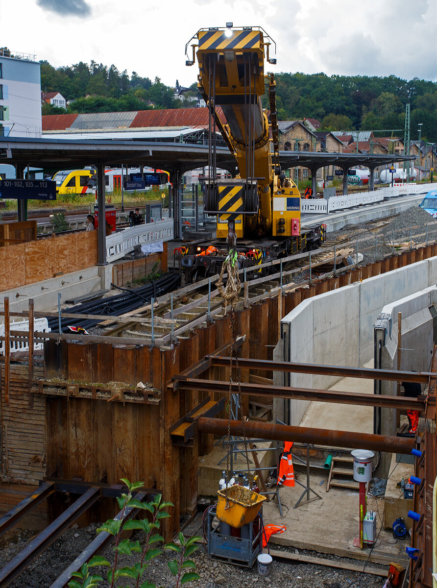 Nun im Einsatz der 125 t KIROW Gleisbauschienenkran KIROW Multi Tasker KRC 810 T (mit Schwerlastausrüstung), Schweres Nebenfahrzeug Nr. D-HGUI 99 80 9419 024-1, ex 97 82 53 513 19-3, der HERING Bahnbau GmbH (Burbach) ist am 29 September 2025 im Betzdorf/Sieg. 

Hier im Bereich unter dem P+R Parkhaus bis zum Fluss Sieg werden Horizontalbohrungen gemacht, vermutlich für erforderliche Entwässerungs-Leitungen. So mussten Bohrrohre in die Baugrube gehoben werden und wie hier der Abraum aus der Baugrube gehoben werden. Eigentlich ist ein 125 t Kran damit unterfordert (überdimensioniert), aber man muss bedenken es wurde eine weite Auskragung (Ausladung vor Puffer) und das freistehend (Nichtabgestützt) benötigt. 

Der hier mit der Ausladung vor Puffer von über 15 m könnte der Kran freistehend 30 t heben, aber darf dann nicht schwenken. Wenn geschenkt werden muss liegt diese dann nur bei 1,5 t. Entscheidend für die Ausladung des Auslegers ist aber auch die hintere Ausladung des Gegengewichts. 