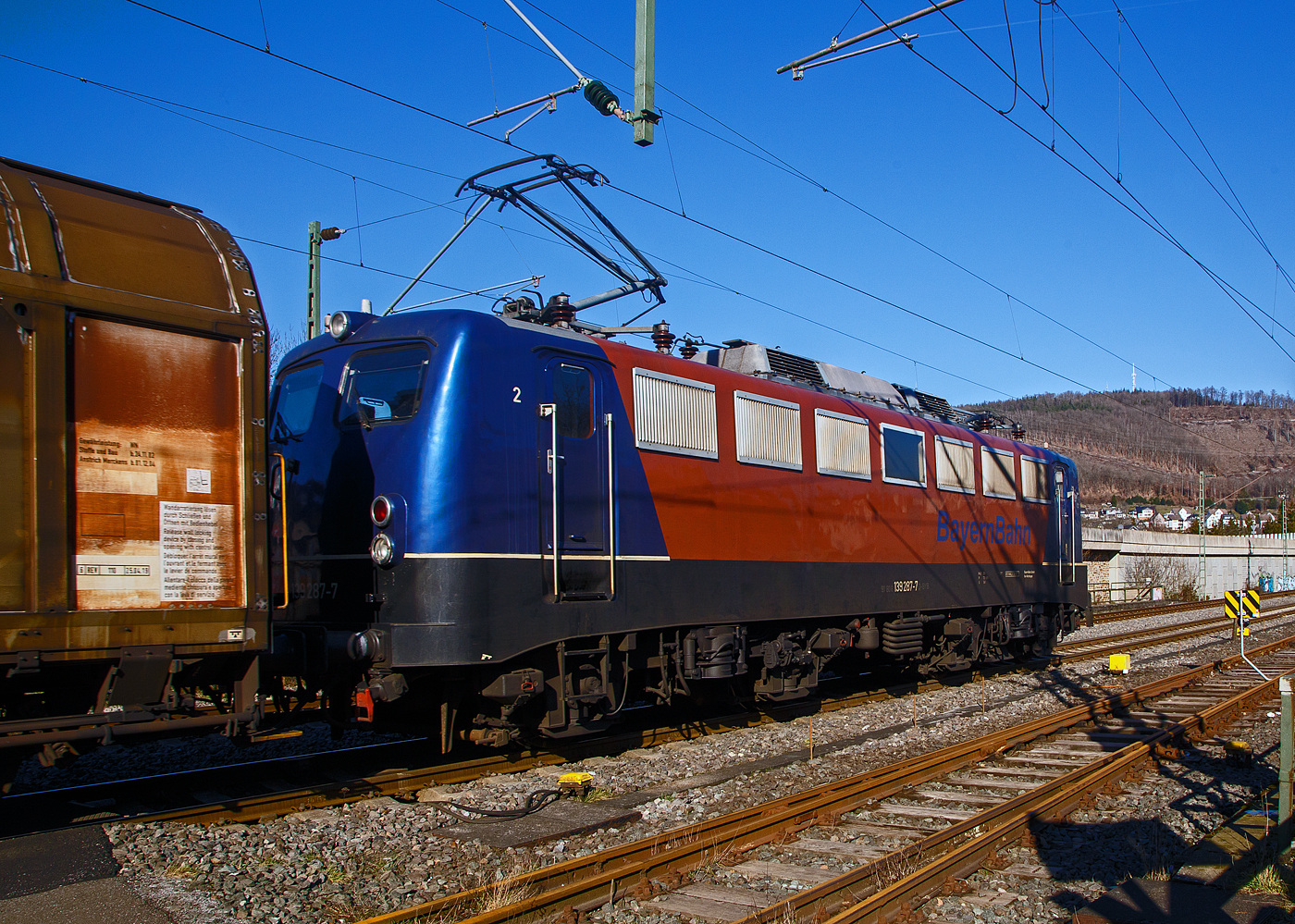 Die 139 287-7 (91 80 6139 287-7 D-BYB) der BayernBahn GmbH fährt am 14.02.2023 mit dem sogenannten  Henkelzug  (Langenfeld/Rhld. nach Gunzenhausen), durch Niederschelderhütte, hier beim Bü 343 (km 112,183) kurz vor Niederschelden bzw. der Landesgrenze zu NRW, in Richtung Siegen.

Nochmal einen lieben Gruß an den netten Lokführer zurück.

Die Lok wurde 1963 von Krauss-Maffei AG in München-Allach unter der Fabriknummer 18956 gebaut, er elektrische Teil ist von den Siemens-Schuckert-Werke (SSW) in Berlin. Als DB E10 287 wurde in Dienst gesetzt, mit der Einführung des EDV-Nummernsystems wurde sie zum 01.01.1968 zur DB 110 287–0. Die Lok ist mit Einholmstromabnehmern ausgerüstet, sie ist die letzte gebaute Kasten 110er. Zur 139er wurde sie erst 1994 nach einem Umbau, der Lokkasten wurde auf Drehgestelle der Baureihe 140 (E40) gesetzt und es folgte die Umzeichnung in DB 139 287-7. Zum Mai 2009 erfolgte die Z-Stellung bei der DB AG. Zum 1.Oktober 2011 wurde die Lok durch die BayernBahn GmbH erworben und erhielt anschließend eine Untersuchung. Die Neulackierung in kobalt-blau/orange erhielt sie dann 2017.
