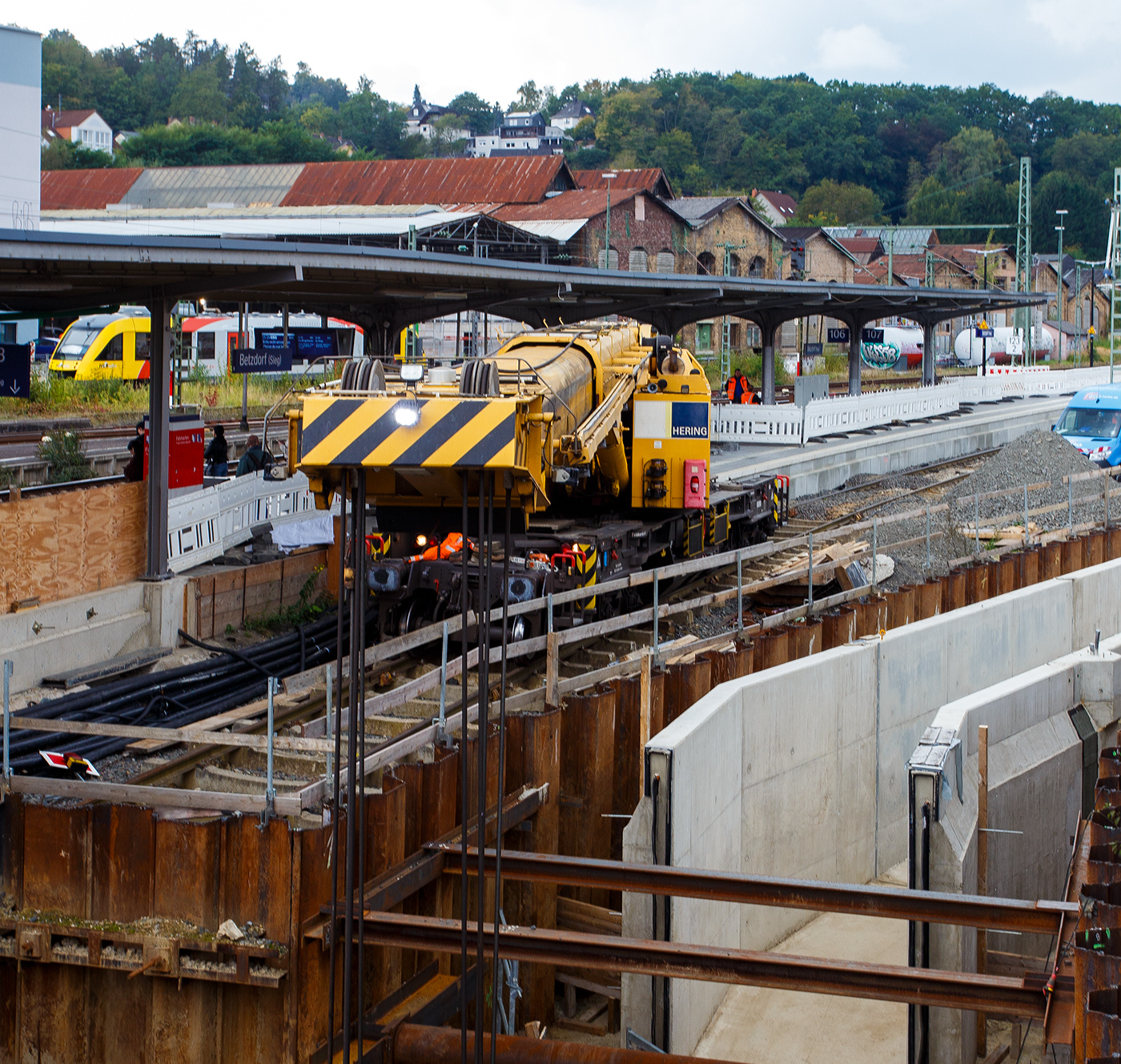 Mal ein Blick über den Teleskopausleger.  
Nun im Einsatz der 125 t KIROW Gleisbauschienenkran KIROW Multi Tasker KRC 810 T (mit Schwerlastausrüstung), Schweres Nebenfahrzeug Nr. D-HGUI 99 80 9419 024-1, ex 97 82 53 513 19-3, der HERING Bahnbau GmbH (Burbach) ist am 29 September 2025 im Betzdorf/Sieg. 

Hier im Bereich unter dem P+R Parkhaus bis zum Fluss Sieg werden Horizontalbohrungen gemacht, vermutlich für erforderliche Entwässerungs-Leitungen. So mussten Bohrrohre in die Baugrube gehoben werden und wie hier der Abraum aus der Baugrube gehoben werden. Eigentlich ist ein 125 t Kran damit unterfordert (überdimensioniert), aber man muss bedenken es wurde eine weite Auskragung (Ausladung vor Puffer) und das freistehend (Nichtabgestützt) benötigt. 

Der hier mit der Ausladung vor Puffer von über 15 m könnte der Kran freistehend 30 t heben, aber darf dann nicht schwenken. Wenn geschenkt werden muss liegt diese dann nur bei 1,5 t. Entscheidend für die Ausladung des Auslegers ist aber auch die hintere Ausladung des Gegengewichts