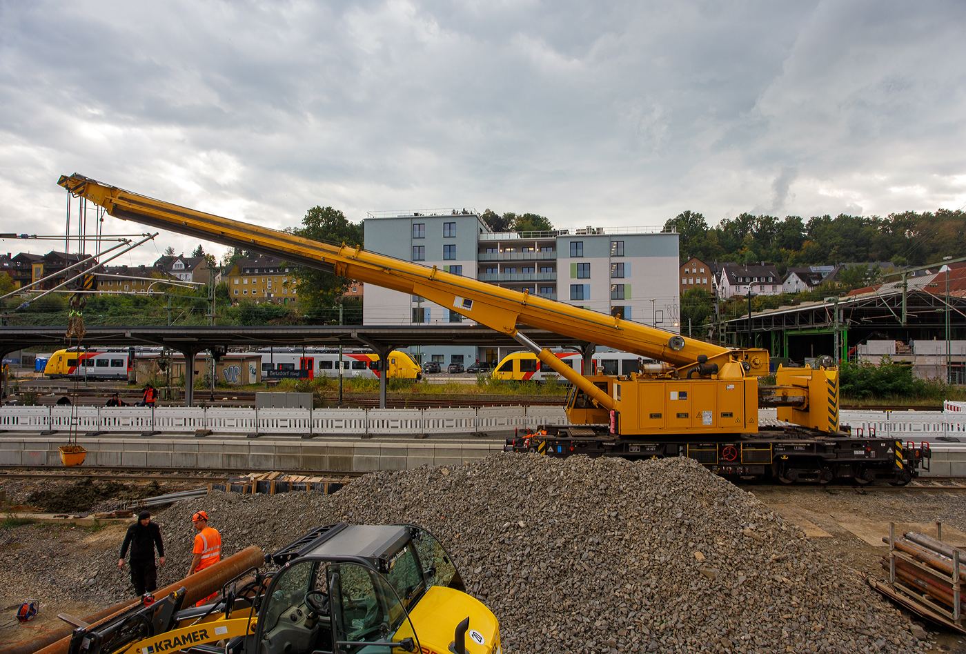 Nun im Einsatz der 125 t KIROW Gleisbauschienenkran KIROW Multi Tasker KRC 810 T (mit Schwerlastausrüstung), Schweres Nebenfahrzeug Nr. D-HGUI 99 80 9419 024-1, ex 97 82 53 513 19-3, der HERING Bahnbau GmbH (Burbach) ist am 29 September 2025 im Betzdorf/Sieg. 

Hier im Bereich unter dem P+R Parkhaus bis zum Fluss Sieg werden Horizontalbohrungen gemacht, vermutlich für erforderliche Entwässerungs-Leitungen. So mussten Bohrrohre in die Baugrube gehoben werden und wie hier der Abraum aus der Baugrube gehoben werden. Eigentlich ist ein 125 t Kran damit unterfordert (überdimensioniert), aber man muss bedenken es wurde eine weite Auskragung (Ausladung vor Puffer) und das freistehend (Nichtabgestützt) benötigt. 

Der hier mit der Ausladung vor Puffer von über 15 m könnte der Kran freistehend 30 t heben, aber darf dann nicht schwenken. Wenn geschenkt werden muss liegt diese dann nur bei 1,5 t. Entscheidend für die Ausladung des Auslegers ist aber auch die hintere Ausladung des Gegengewichts. 

Der Kran wurde 2008 von KIROW in Leipzig unter der Fabriknummer 149000 gebaut und an die HERING Bahnbau GmbH (Burbach) geliefert.

Der Gleisbauschienenkran KRC 810 T ist ein Schienendrehkran des Herstellers Kirow Ardelt (bis 2008 Kirow) in Leipzig. Der Typ 810 wird für den Aus-und Einbau von schweren Betonweichen, Gleisjochen und Hilfsbrücken sowie für die Montage von Ingenieurbauwerken eingesetzt. Bei Havarien auf Bahnstrecken leistet er schnelle Hilfe zum Bergen schwerer Lasten. Das maximale Lastmoment des Krans liegt zwischen 500 und 900 tm. Die Krane werden auch als Eisenbahndrehkran „EDK“ bezeichnet.

Die maximale Tragfähigkeit des bis zu 128 Tonnen schweren Krans beträgt abgestützt bis zu 125 Tonnen und freistehend 90 Tonnen. Bei maximaler Ausladung kann der Kran noch 28,5 Tonnen heben. Die größte mögliche Hakenhöhe liegt bei 18 m und die maximale Ausladung vor Puffer bei 15 m. Der Teleskoparm erlaubt Arbeiten im Radius zwischen 6,5 und 24,5 Metern. Im Gegensatz zu früheren Kranen ist auch ein Einsatz in der Nullposition ohne Abbau der Fahrleitung möglich. Die Gegenlast (Gegengewicht) ist zum Ausgleich unterschiedlicher Lasten ausfahrbar. Die Gegenlast kann in Transportstellung nicht (wie z.B. bei dem KRC 910) auf einem Gegenlastwagen abgestützt werden. Dadurch hat KRC 810 auch eine höhere Streckenlast (als z.B. der KRC 910). Für Hebearbeiten instehender Stellung verfügt der Kran über hydraulisch-ausfahrbare Stützen. Die Höchstgeschwindigkeit 19 km/h mit dem Eigenantrieb und 120 km/h im Zugverband geschleppt.  Zu dem Kran gehören zwei Kranschutzwagen. Die zulässige Anhängelast beträgt 80 t.

TECHNISCHE DATEN:
Spurweite: 1.435 mm (Normalspur)
Anzahl der Achsen: 8
Achsfolge: [(1A)(1A)][(A1)(A1)]
Länge über Puffer: 13.000 mm (ohne Ausleger)
Drehzapfenabstand: 8.000 mm
Drehzapfenabstand im Drehgestell 1 und 2: 2.300 mm
Achsabstand in den Einzeldrehgestellen: 1.100 mm
Ergebene Achsabstände in m: 1,1 / 1,2 / 1,1/ 4,6 / 1,1 / 1,2 / 1,1
Treib- und Laufraddurchmesser: 730 mm (neu) / 680 mm (abgenutzt)
Höhe in Transportstellung: 4.250 mm
Breite in Transportstellung: 3.100 mm
Eigengewicht: 128 t
Dieselmotor: Wassergekühlter 6-Zylinder Reihenmotor mit Abgasturboaufladung und Ladeluftkühlung vom Typ Cummins M 11 – C 315
Motorleistung: 235 kW bei 1.800 U/min
Max. Drehmoment: 1.458 Nm bei 1.300 U/min
Not-Dieselmotor: Luftgekühlter 2-Zylinder Viertakt-Dieselmotor mit Direkt-Einspritzung vom Typ Hatz 2 G 40 mit einer Leistung11,5 kW bei 2.300 U/min.
Fahrgeschwindigkeit im Zugverband: 120 km/h (geschleppt)
Fahrgeschwindigkeit mit Eigenantrieb: 20 km/h
zul. Anhängelast: 80 t
Hakenhöhe (max.): 18 m
Ausladung (max. vor Puffer): 16 m
Zugelassen als Schwerwagen für Streckenklasse: CS 05
Maximale Radsatzlast: 16 t
Streckenlast (Meterlast):  9,9 t/m
kleinster befahrbarer Radius: 00 m
Bremse: KE-GP-D- m.Z. (Scheibenbremsen auf allen Rädern)
Sekundär Scheibenbremsen auf allen Rädern
Handbremse: hydro-mechanisch auf alle Radsätze
Kraftstoffvorrat: 990 l

Tragfähigkeiten:
bei 0,00 m Ausladung vor Puffer, mit Schwerlastausrüstung: 125 t
bei 2,80 m Ausladung vor Puffer, ± 30°, abgestützt: 90 t
bei 2,80 m Ausladung vor Puffer, ± 20°, freistehend: 46 t