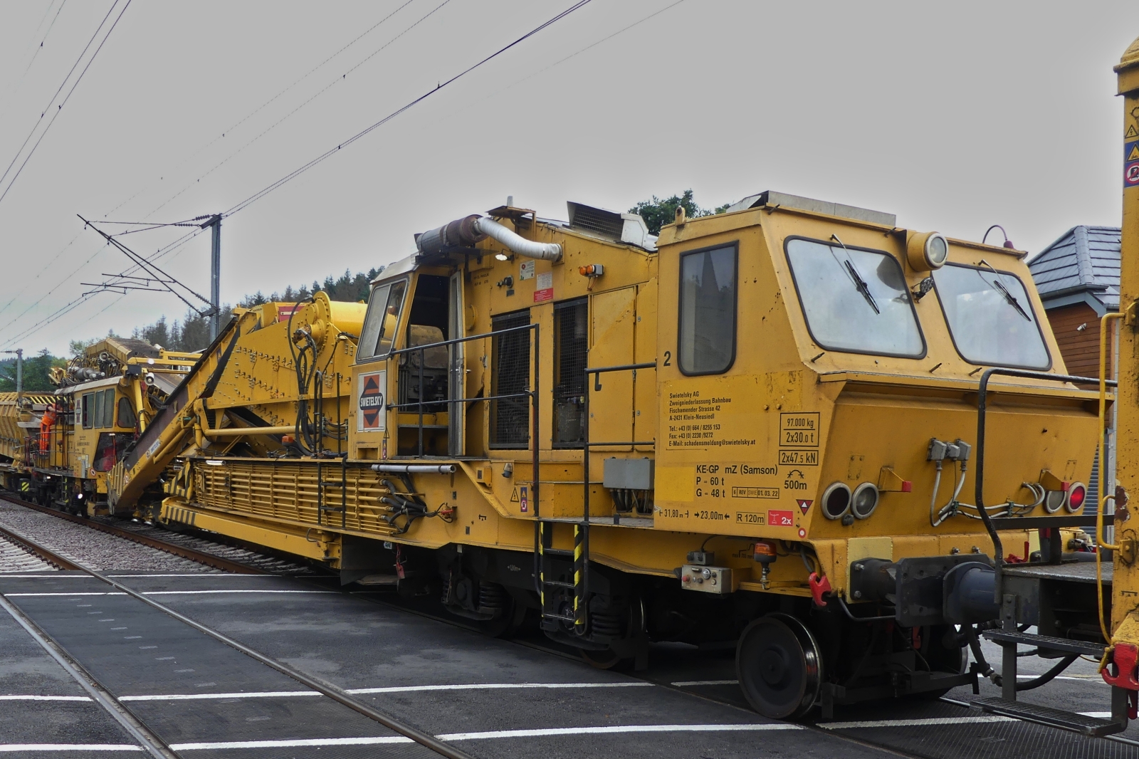 Swietelsky Schotterbettreinigungsmaschine RM 80-92 (9981 9414 001-3) schiebt die Schottersammelwagen im Schneckentempo über den Bahnübergang in Enscherange. 25.08.2023