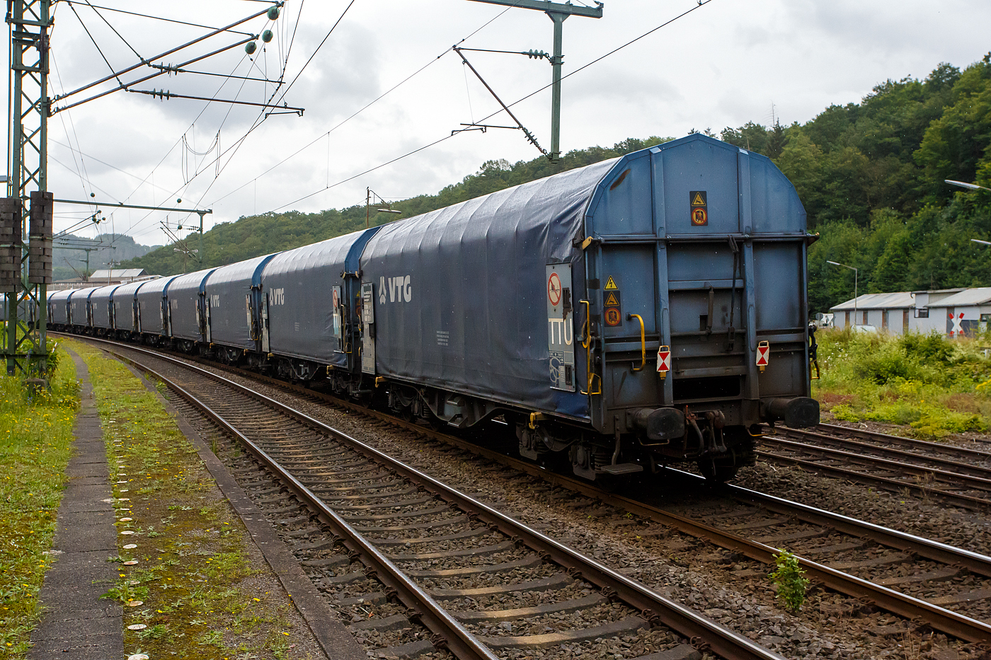 Und tschüss bis gleich in Herdorf, der vierachsiger Drehgestell-Flachwagen mit verschiebbarem Planenverdeck und Lademulden für Coiltransporte, 37 80 4668 122-7 D-VTG, der Gattung Shimmns-ttu (VTG Bez. F46.520D), der VTG Rail Europe GmbH (Hamburg), am 03 Juli 2024 eigereiht im Zugverband (Zudschluss) der KSW bei einer in Zugdurchfahrt im Bf Scheuerfeld/Sieg, in Richtung Herdorf via Betzdorf.

Diese Wagen sind speziell für den Transport witterungsempfindlicher Blechrollen (Coils) bestimmt. Er besitzt feste Stirnwände, 5 fest im Untergestell eingebaute Lademulden. Die Wagenabdeckung erfolgt mit einer PVC-beschichteten Gewebeplane, die auf 11 Rohrrahmen aufliegt. Über Laufwagen ist das Planendach in Wagenlängsrichtung verschiebbar. Die Verriegelung des Planendaches an den Stirnwänden erfolgt über eine 4-Punkt-Zentralverriegelung, die sowohl vom Erdboden als auch von der Rampe aus bedient werden kann. Das Planenverdeck lässt sich an einem Wagenende so weit zusammenschieben, dass ca. 2/3 der Ladefläche zur Beladung freigegeben wird. Hierdurch ist eine einfache Beladung des Wagens sowohl von oben als auch von der Seite aus möglich.

TECHNISCHE DATEN:
Spurweite: 1.435 mm
Achsanzahl: 4 (in 2 Drehgestelle)
Länge über Puffer: 12.040 mm
Länge der Ladefläche: 10.800 mm
Drehzapfenabstand: 7.000 mm
Achsabstand im Drehgestell: 1.800 mm
Eigengewicht: 22.250 kg
Höchstgeschwindigkeit: 100 km/h (beladen) / 120 km/h (leer)
Maximales Ladegewicht: 67,7 t (ab Streckenklasse D4), DB CM 61,7 t
Mulden: 5 Stück, im Untergestell integriert
Werkstoff Muldenmatten: Tecthan
Festlegearme: 20 Stück, stufenlos verstellbar, ausgestattet mit Schutzplatten
Coil-Breite: min. 342 mm bis max.  2.042 mm
Kleinster bef. Gleisbogenradius: 35 m
Bauart der Bremse: KE-GP-A (K)
Bremssohle: C 810
Intern. Verwendungsfähigkeit: TEN-GE