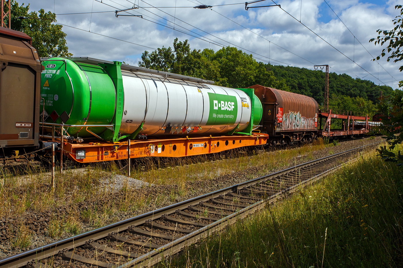Vierachsiger Containertragwagen 33 85 4505 278-3 CH-WASCO der Gattung Sgmmns 54‘, für den Transport von intermodalen Ladeeinheiten und des Wascosa flex freight system (Wechselaufbauten), der Wascosa AG am 11 Juli 2025 im Zugverbund bei einer Zugdurchfahrt in Rudersdorf (Kreis Siegen). Der Wagen ist beladen, mit einem vom belgischen Hersteller Van Holl gebauten, 52 ft Tankcontainer der BASF, der hier laut Gefahrguttafel 83/2734 mit Amine oder Polyamine befüllt ist. Die Kombination des Wagens mit 45‘ oder 52‘ Tankcontainern kann Kesselwagenersetzen und ermöglicht eine unabhängige Instandhaltung beider Komponenten.

Gebaut wurde der Wagen vom slowakischen Güterwagenhersteller Tatravagónka in Poprad.

TECHNISCHE DATEN (Wagen):
Spurweite: 1.435 mm (Normalspur)
Länge über Puffer: 17.800 mm
Drehzapfenabstand: 11.920 mm
Radsatzstand in den Drehgestellen: 1.800 mm
Ladelänge: 16.470 mm
Höhe der Ladeebene für Container über S.O.: 1.105 mm
Drehgestell: Y25 Lsi(f)-C-K
Raddurchmesser: 920 mm (neu) / 860 mm (abgenutzt
Eigengewicht: 16.500 kg
Max. Zuladung bei Lastgrenze S: 73,5 t (ab Streckenklasse D)
Max. Geschwindigkeit: 100 km/h (leer 120 km/h)
Max. Radsatzlast: 22,5 t
Druckluftbremse: Knorr KE-GP-A (K)
Bremssohle: C 810
Handbremse: Ja
Kleinster bef. Gleisbogenradius: 150 m (im Zugverband) / 75 m der Einzelwagen
Intern. Verwendungsfähigkeit: TEN GE

Der Tankcontainer:
Der 52-Fuß-Tankcontainer mit 75 Tonnen zulässigem Gewicht (bei einem Eigengewicht von 8.200 kg) und 73.500 Liter Inhalt, hat die doppelte Ladekapazität wie ein heute üblicher Tankcontainer und eine vergleichbare Kapazität wie ein Chemiekesselwagen. Der neue, bahnoptimierte Tankcontainer hat die gleiche Technik wie die bisher üblichen 20 bis 30 Fuß messenden Tankcontainer. Die 52 Fuß-Tankcontainer können zum Bahntransport, Binnenschiffstransport und, im leeren Zustand, auch zum Straßentransport eingesetzt werden. Darüber hinaus sind sie zur Lagerung in Containerlagern zugelassen. Ausgestattet sind sie außerdem mit moderner Isoliertechnik sowie Heizvorrichtungen. Der 52 Fuß-Tankcontainer verfügt im Vergleich zum Bahnkesselwagen über ein höheres Ladegewicht im Verhältnis zum Eigengewicht. Dank der Möglichkeit, vom Bahnwagen abgenommen werden zu können, und der Stapelbarkeit benutzt der Tankcontainer deutlich weniger Infrastruktur und ist flexibler beim Be- und Entladen.

Entwickelt wurden die 45 und 52 Fuß Tankcontainer für den Bahnverkehr von Van Hool und BASF gemeinsam. Der erste Prototyp wurde 2015 geliefert.